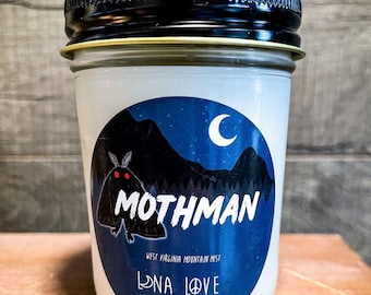 May include: A white candle jar with a blue and black label featuring a mothman illustration, a crescent moon, and the text "Mothman", "West Virginia Mountain Mist", "Luna Love Candles".