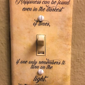 May include: A rectangular light switch cover with a beige, parchment-like finish. It features black cursive text that reads: Happiness can be found even in the darkest of times, if one only remembers to turn on the light. -Albus Dumbledore.