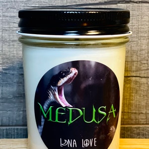 Puede incluir: Un tarro de vela blanco con tapa negra y una etiqueta que dice "Medusa" en letras verdes. La etiqueta también presenta una imagen en blanco y negro de una serpiente con la boca abierta y un signo de paz. La etiqueta también dice "Luna Love Candles".