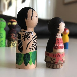 May include: Wooden peg dolls, hand-painted with detailed designs. One doll features black hair and a tan body with black tribal tattoos and green leaves. The other doll has black hair and a red and white patterned top. Other dolls are visible in the background.