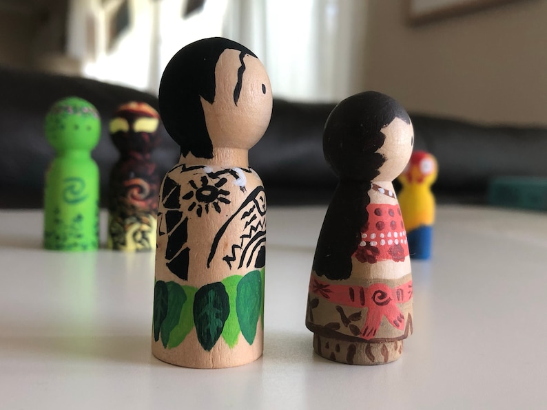 May include: Wooden peg dolls, hand-painted with detailed designs. One doll features black hair and a tan body with black tribal tattoos and green leaves. The other doll has black hair and a red and white patterned top. Other dolls are visible in the background.