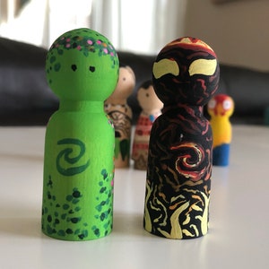 May include: Two wooden peg dolls, one green with floral and wave designs, the other black with flame and swirl patterns. Other dolls are visible in the background. The dolls are hand-painted.