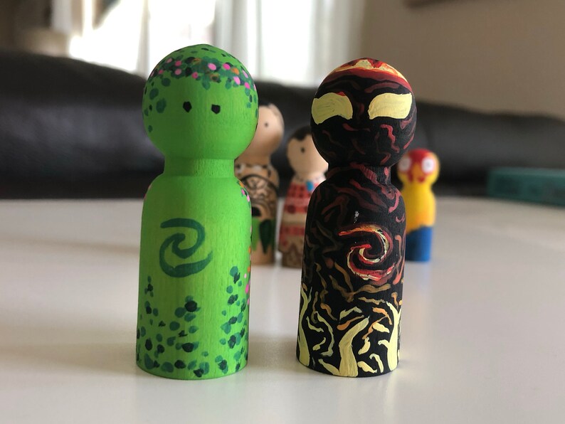 May include: Two wooden peg dolls, one green with floral and wave designs, the other black with flame and swirl patterns. Other dolls are visible in the background. The dolls are hand-painted.