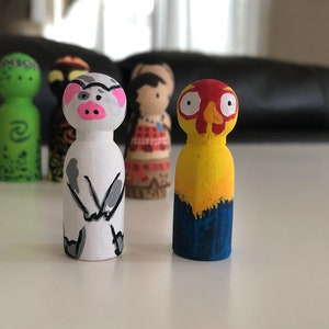 May include: A collection of hand-painted wooden peg dolls. The dolls feature various character designs, including a pig with a pink snout, a yellow and red bird-like figure, and other figures with detailed facial features and clothing.