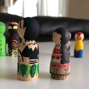 May include: Wooden peg dolls, hand-painted with colorful designs. One doll features a black and tan design, while another has a red and white pattern. Other dolls in the background have green, yellow, and orange paint.
