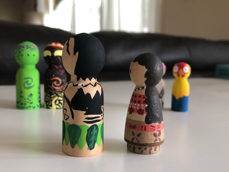 May include: Wooden peg dolls, hand-painted with colorful designs. One doll features a black and tan design, while another has a red and white pattern. Other dolls in the background have green, yellow, and orange paint.