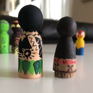 May include: Wooden peg dolls, hand-painted with black, green, and brown details. One doll has a black head and a design of black shapes and green leaves. The other doll has a black head and a design of orange and brown floral patterns.