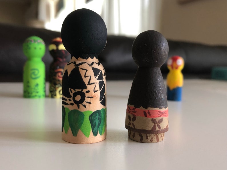 May include: Wooden peg dolls, hand-painted with black, green, and brown details. One doll has a black head and a design of black shapes and green leaves. The other doll has a black head and a design of orange and brown floral patterns.