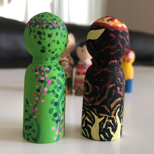 May include: Two hand-painted wooden peg dolls. One is green with pink and orange floral details. The other is black with orange and yellow flame-like designs. Other dolls are visible in the background.