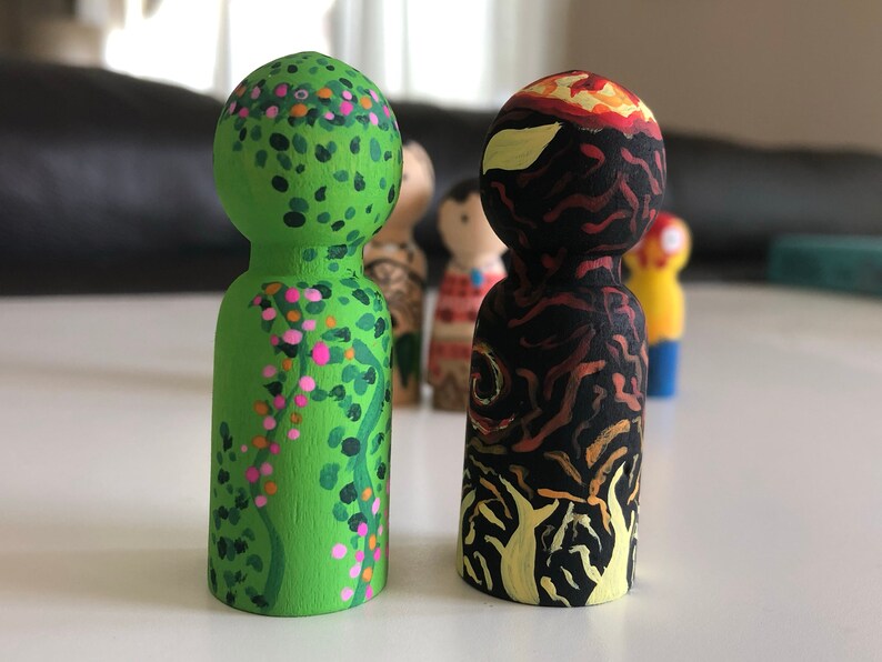 May include: Two hand-painted wooden peg dolls. One is green with pink and orange floral details. The other is black with orange and yellow flame-like designs. Other dolls are visible in the background.