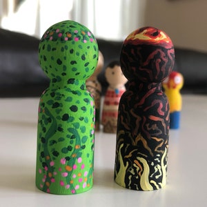 May include: Two wooden peg dolls, one green with floral details and the other black and red, depicting a volcano. The dolls are hand-painted with intricate designs and stand upright. Other dolls are visible in the background.