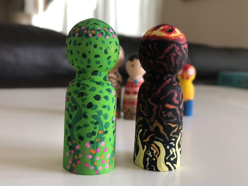 May include: Two wooden peg dolls, one green with floral details and the other black and red, depicting a volcano. The dolls are hand-painted with intricate designs and stand upright. Other dolls are visible in the background.