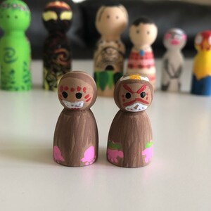 May include: Hand-painted wooden peg dolls with various character designs. Two dolls in the foreground feature brown bodies and faces with red and white details. Other dolls in the background have different colors and designs, including green, yellow, and red.