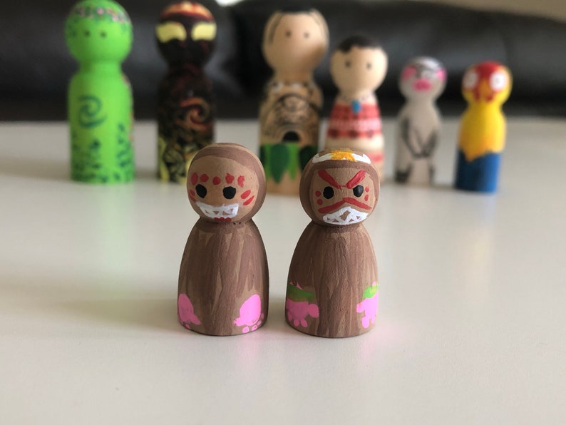May include: Hand-painted wooden peg dolls with various character designs. Two dolls in the foreground feature brown bodies and faces with red and white details. Other dolls in the background have different colors and designs, including green, yellow, and red.