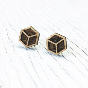 May include: A pair of wooden stud earrings. Each earring is a hexagon shape with a cube design etched into the wood. The earrings are a light brown color with a darker brown cube design. The background is a white, distressed wood surface.