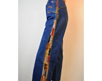 May include: High-waisted blue denim jeans featuring a wide, vertical side stripe composed of a multicolored, rectangular patchwork pattern in shades of red, orange, yellow, white, and black, extending from the waistband down the leg.