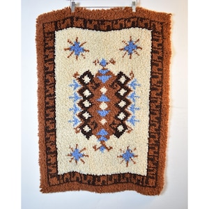May include: A rectangular, textured wall hanging with a cream-colored center, brown border, and brown fringe. The center features geometric patterns in brown, blue, and white, including star-like shapes. The wall hanging is suspended from a metal hanger.