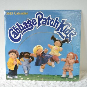 May include: A 1985 Cabbage Patch Kids calendar featuring a blue cover with five dolls playing jump rope on a green lawn. The dolls wear various outfits including a yellow jacket, blue overalls, a plaid dress, a yellow shirt, and a peach dress.