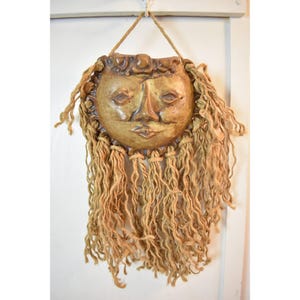 May include: A ceramic sun face wall hanging with a detailed face and long, textured hair made of rope. The sun face is a warm, earthy tone, and the rope hair is a light brown color. The piece is suspended by a natural fiber cord.