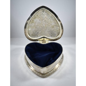 May include: A heart-shaped metal trinket box with a silver-toned exterior and a hinged lid. The interior of the box is lined with deep blue velvet fabric. The underside of the lid features a textured, reflective silver surface with a mottled pattern.