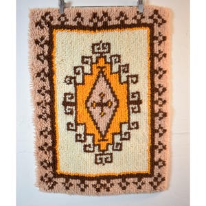 May include: A vintage, rectangular wall hanging with a geometric design. The central pattern features a diamond shape in orange and pink, surrounded by brown and cream-colored elements. The border is a soft pink with a textured, fringed edge.