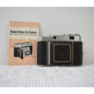 May include: A vintage Kodak Retina IIa camera with a black and silver body, positioned next to its original instruction manual. The manual features the text Kodak Retina IIa Camera, Retina-Xenon f/2 Lens, and Synchro-Compur Shutter.