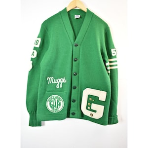 May include: Green vintage cardigan sweater with a V-neck and button closure. Features white patches with the letters "G", "D", and "A", and the word "Muggs". The sleeve has a white stripe with the number "5". The bottom patch reads "Greenway High".