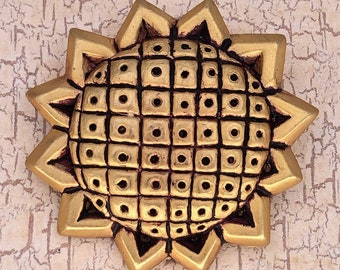 May include: A gold-toned metal brooch in the shape of a stylized sun with a textured center. The center is a grid of small squares with a circle in each square.