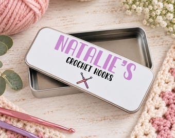 Puede incluir: Una caja rectangular de metal plateado con una tapa blanca que presenta el texto morado NATALIE'S y el texto negro CROCHET HOOKS, acompañado de un pequeño gráfico de dos ganchillos rosas cruzados.