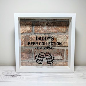 May include: A square white shadow box frame featuring a brick wall background. The front glass displays black text reading DADDY'S BEER COLLECTION - Est.2024 - above a graphic of two clinking beer mugs. The frame has a slot at the top for inserts.