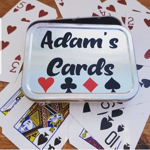 May include: A silver metal card case with the text "Adam's Cards" printed on the front. The case is surrounded by playing cards.