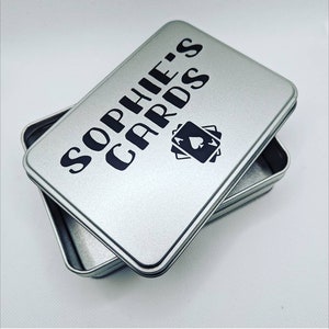 May include: A silver metal box with a black inscription that reads "Sophie's Cards" and a black image of playing cards with an ace of spades.
