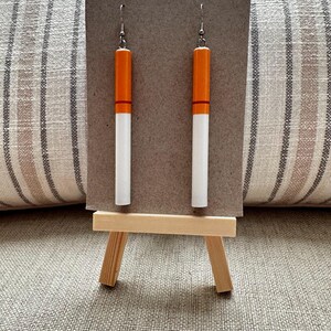 Op de afbeelding: Een paar opvallende oorbellen die op sigaretten lijken. Elke oorbel heeft een witte cilindrische behuizing met een oranje punt en een rode band, opgehangen aan een zilveren haak. De oorbellen worden op een klein ezelbord gepresenteerd.