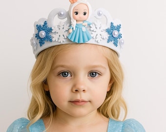 May include: A young girl wearing a light blue dress and a white crown with snowflake and doll embellishments. The dress has a snowflake applique and sheer sleeves. The crown features a small doll figure.