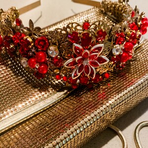 May include: A gold-coloured metal headband adorned with red and clear gemstones, alongside a gold clutch with a chain strap. The headband showcases a floral design with red gems and clear crystals. The clutch has a mesh-like texture.