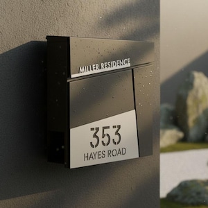 May include: A modern black mailbox mounted on a gray wall. The mailbox features the text "MILLER RESIDENCE" and the address "353 HAYES ROAD" in a clean, minimalist font. The design incorporates a diagonal silver accent.