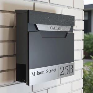 May include: A modern, dark gray mailbox mounted on a white brick wall. The mailbox features a silver nameplate with the name "CALLAS" and a lower silver panel with the address "Milson Street 25B".