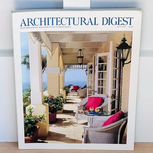 Może przedstawiać: Okładka magazynu Architectural Digest z lipca '91. Okładka przedstawia słoneczną scenę na zewnątrz z werandą, wiklinowymi meblami i widokiem na ocean. Tytuł magazynu jest w dużych, niebieskich literach.