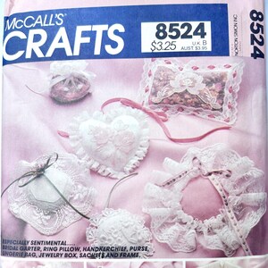 May include: A McCall's Crafts 8524 sewing pattern envelope featuring a pink background with images of white lace bridal accessories including a garter, ring pillow, handkerchief, purse, lingerie bag, jewelry box, sachets, and a picture frame.