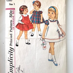 Peut inclure: Une pochette de patron de couture vintage Simplicity, numéro 5165, pour vêtements de tout-petit taille 1. Le devant présente des illustrations de trois enfants portant différents styles de robes, dont une chasuble rouge, une robe à carreaux et une robe blanche avec une girafe.