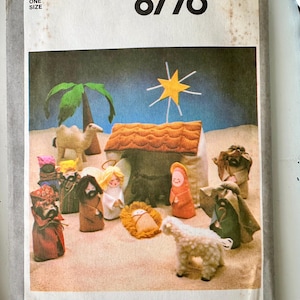 May include: A Simplicity 8776 sewing pattern envelope featuring a nativity scene illustration. The image displays fabric figures including a camel, a stable, a star, a sheep, and various characters surrounding a manger on a sandy surface.