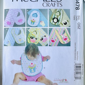 May include: McCall's Crafts M6478 sewing pattern envelope for baby bibs. The front displays six variations labeled A through F, featuring animal and cupcake appliqués on patterned fabrics. A child wears bib B, a white bib with colorful dots.