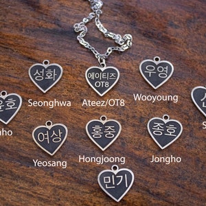 Könnte beinhalten: Eine silberne Halskette mit acht herzförmigen Anhängern, jeder mit einem anderen koreanischen Namen in Schwarz geschrieben. Die Namen sind Seonghwa, Wooyoung, Yunho, Yeosang, Hongjoong, Jongho, San und Mingi. Die Halskette hat auch einen Anhänger mit dem Text "Ateez/OT8".