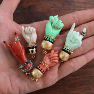 Könnte beinhalten: Eine Sammlung von fünf Miniatur-Handskulpturen in verschiedenen Farben, darunter Rot, Weiß, Grün und Pfirsich. Jede Hand ist mit goldfarbenen Armbändern und Ringen verziert. Die Hände sind in verschiedenen Gesten, wie dem 'Ich liebe dich'-Zeichen und dem 'Rock on'-Zeichen.