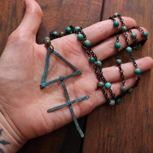 May include: A handcrafted necklace featuring a pendant with a geometric design and a chain adorned with turquoise beads. The pendant has a weathered, teal patina. The necklace is displayed on a wooden surface.