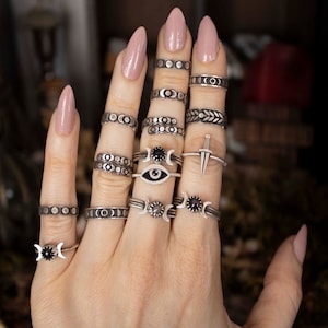 Può includere: Una collezione di anelli d'argento con vari disegni, tra cui lune crescenti, cerchi, un occhio e una piccola spada. Alcuni anelli presentano pietre nere. Gli anelli sono esposti su una mano con unghie lunghe e smaltate.