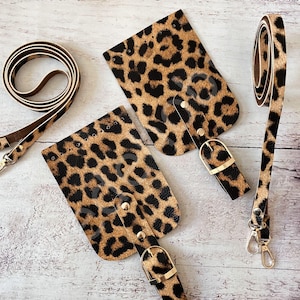 Puede incluir: Dos fundas para teléfono de cuero con estampado de leopardo marrón y negro con hebillas y correas doradas. Las correas están unidas a las fundas con remaches dorados.