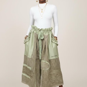 May include: A woman wearing a white long-sleeve top and olive green culottes. The culottes have a high waist, pockets, and a textured, patchwork design. The outfit is accessorized with gold jewelry and nude heels.