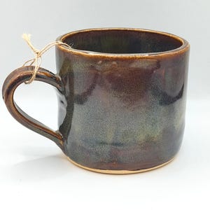 May include: A ceramic mug with a glossy, multi-toned glaze featuring deep brown, charcoal, and subtle green hues. The mug has a rounded handle with a thin, light-colored twine string tied around it. The base of the mug shows the natural clay color.