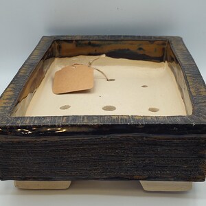 May include: A rectangular ceramic bonsai pot with a dark brown, textured exterior and a smooth, cream-colored interior. The pot features a wide rim, four small feet, and several drainage holes in the base, with a blank brown paper tag attached.